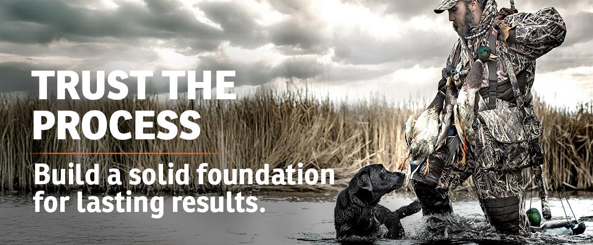Hunter wading through water with his black lab walking next to him and looking up at him with text: Trust the Process. Build a solid foundation for lasting results.
